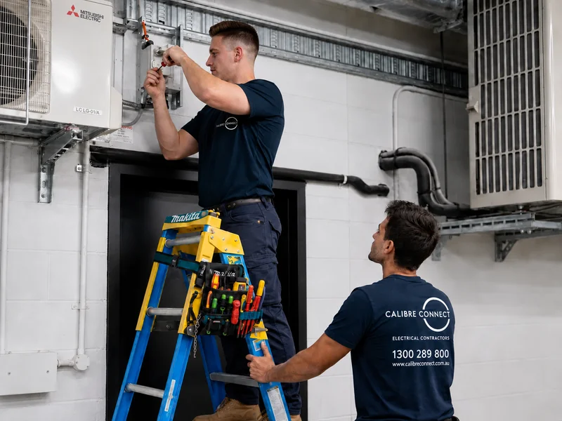 Two Calibre Connect electricians working from a ladder on equipment circuits during RCD safety switch testing in Sydney